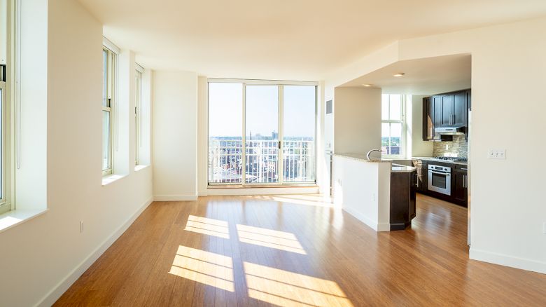 This image shows a bright, empty apartment with large windows, wooden floors, an open kitchen, and a view of the city from the balcony.