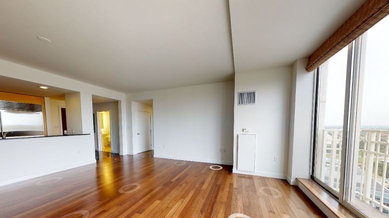 A modern, unfurnished living room with wooden flooring, large windows, and a view of the cityscape, adjacent to an open kitchen space.