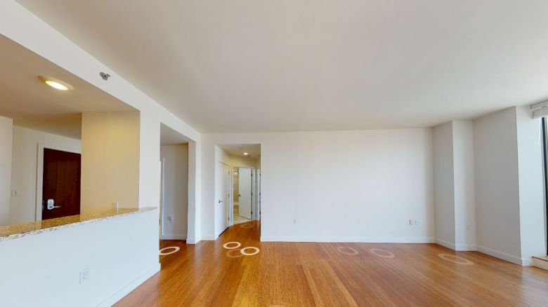 The image shows a modern, empty apartment interior with wooden floors, white walls, a kitchen area to the left, and large windows to the right.