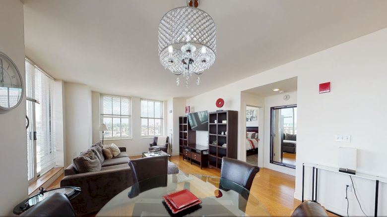 A modern living room with dining area, featuring a chandelier, TV, bookshelves, and a view into a bedroom. Light-filled space with wooden floors.