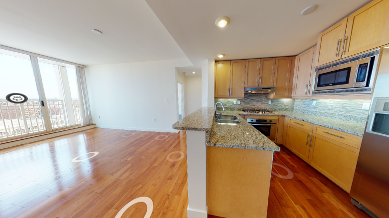 The image shows a spacious kitchen-living area with hardwood floors, granite countertops, and stainless steel appliances, including a fridge and microwave.