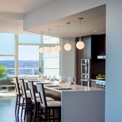 A modern kitchen features a white island with chairs, pendant lights, stainless steel appliances, and a view of large windows and the outdoors.