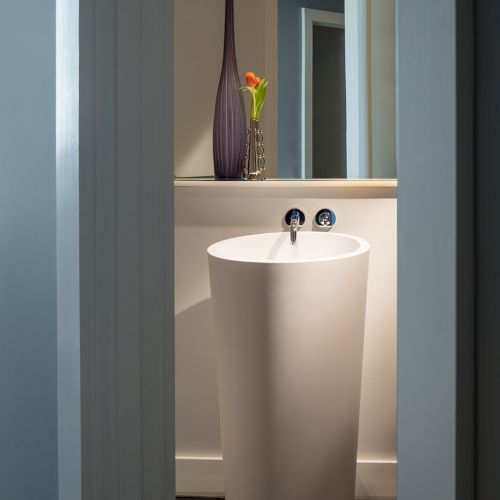 A modern bathroom sink with a tall, cylindrical design is shown in a minimalistic space, accompanied by a vase with a flower arrangement.