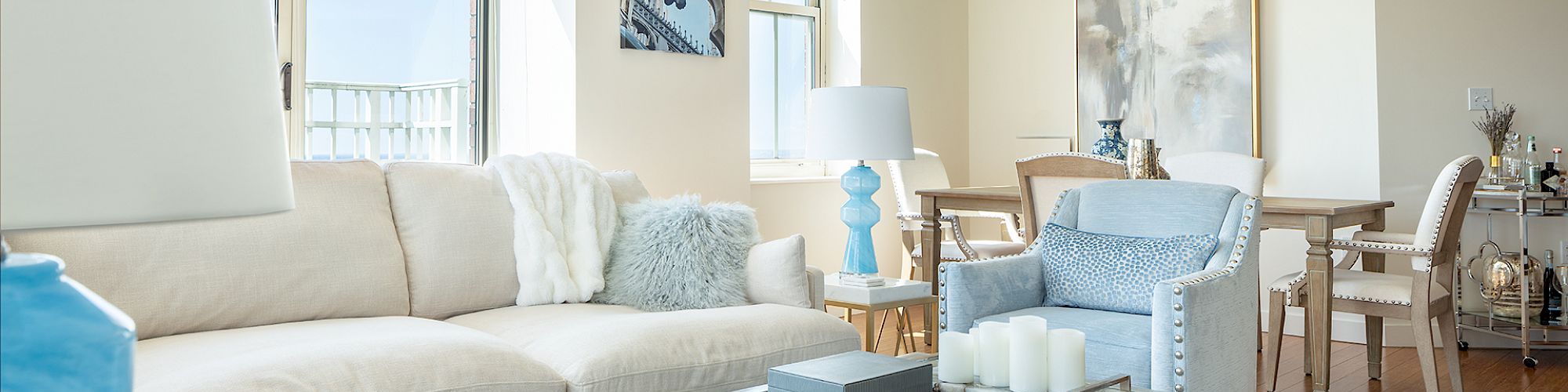 A well-lit living room with modern décor, white sofa, glass coffee table with books and candles, blue accents, dining area with chandelier in the background.