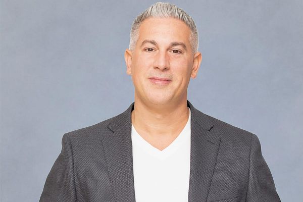 The image shows a man with short gray hair wearing a gray blazer over a white shirt, standing against a plain gray background.