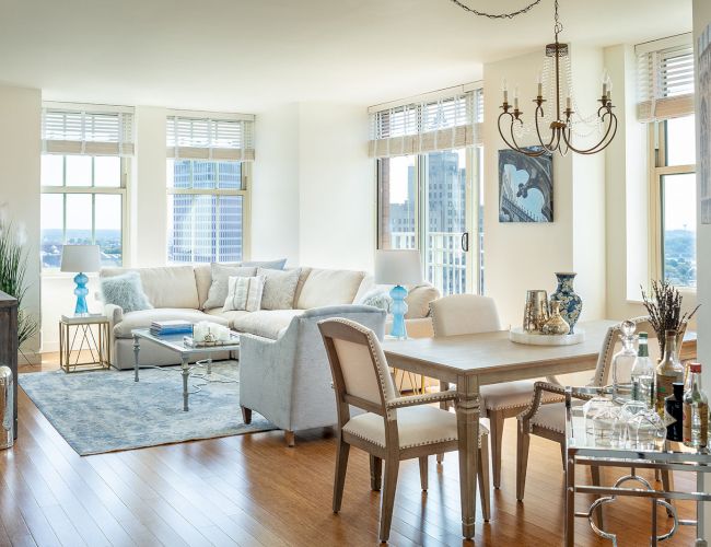 A modern, bright living and dining area with large windows, a white sofa, a dining table, a bar cart, and chic decor accents, offering skyline views.